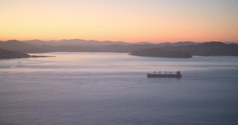 A large cargo ship sails through a calm bay at sunset with rolling, hazy mountains in the background