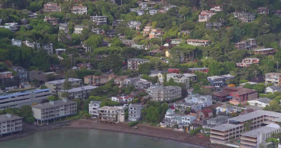 An aerial shot slowly pans across the picturesque coastal town of Sausalito, California, revealing beautiful homes built into the lush, green hillside along the bay