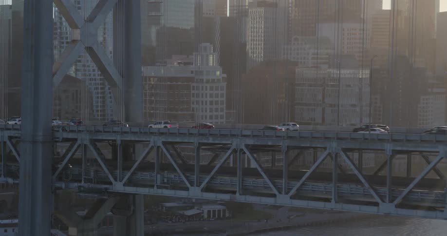 An aerial view of traffic moving along the San Francisco-Oakland Bay Bridge with the city skyline in the background at sunrise