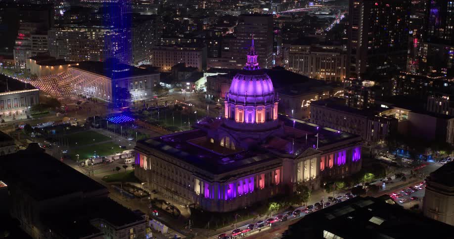 A slow aerial pullback reveals the magnificent San Francisco City Hall illuminated in vibrant purple at night, set against the backdrop of the bustling city and bay