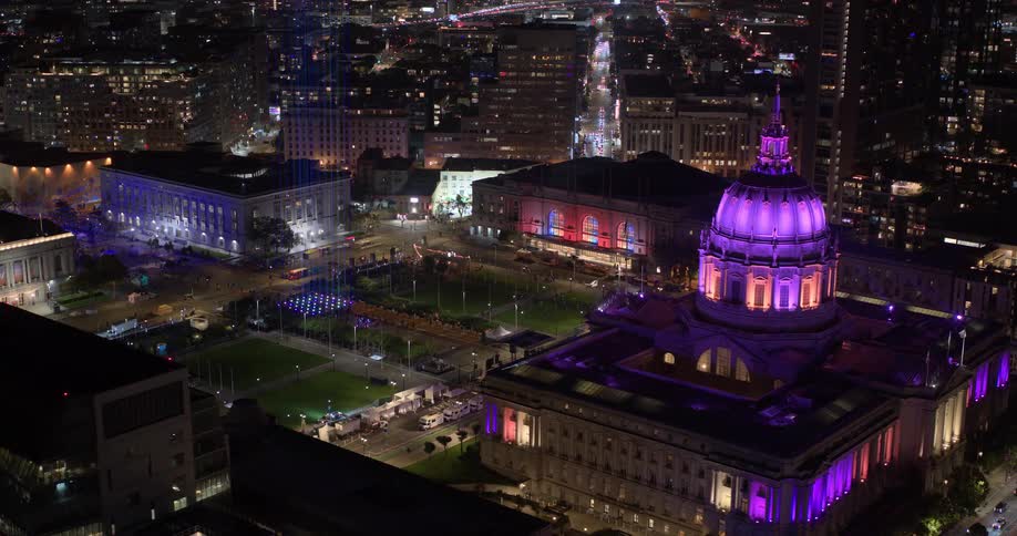 A stunning aerial view shows the San Francisco City Hall illuminated in vibrant purple lights, surrounded by the bustling city at night