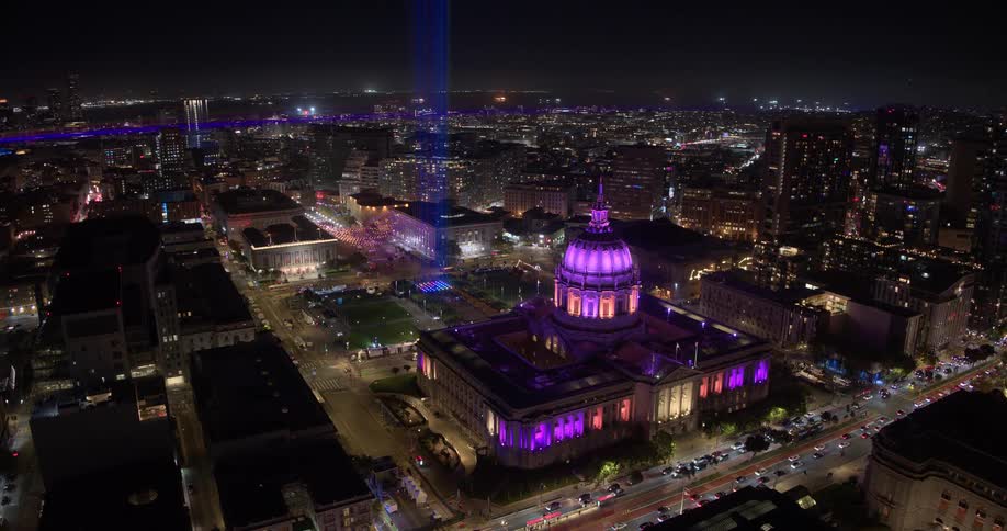 Cinematic aerial time-lapse of the brightly illuminated San Francisco City Hall at night, with vibrant city lights and traffic trails filling the surrounding streets