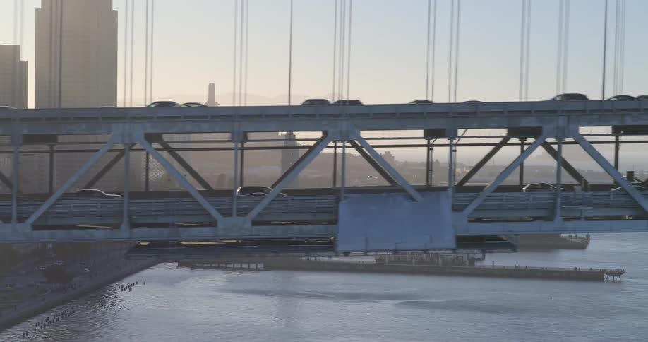 An aerial side view of traffic flowing across the double-decker San Francisco-Oakland Bay Bridge at sunrise with the city skyline in the background