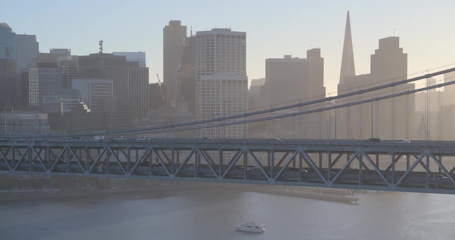A slow pull-back aerial view reveals traffic crossing the Bay Bridge with the hazy San Francisco skyline glowing in the background at sunrise