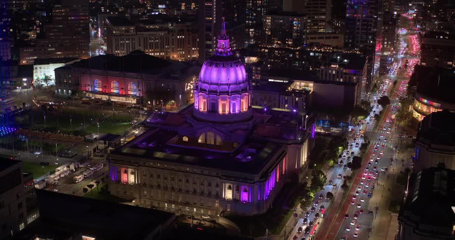 A slow zoom-out timelapse captures the iconic San Francisco City Hall illuminated in vibrant purple as traffic light trails streak through the bustling cityscape at night