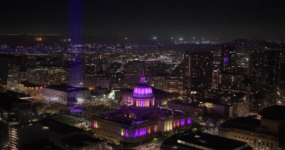 A slow aerial pull-back reveals the majestic San Francisco City Hall and surrounding cityscape, beautifully illuminated with purple and blue lights at night