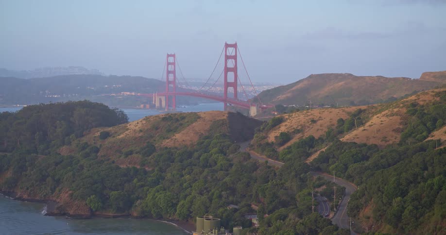 A wide, static shot of the iconic Golden Gate Bridge from the Marin Headlands with a winding coastal road in the foreground at dusk