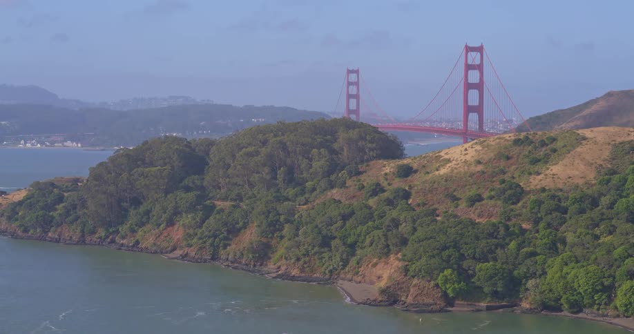 Aerial view pans across the Marin Headlands to reveal the iconic Golden Gate Bridge over the San Francisco Bay on a hazy day