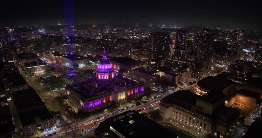 Stunning aerial time-lapse of San Francisco City Hall and Civic Center illuminated in purple at night, with a light beam shooting into the sky as traffic streaks by