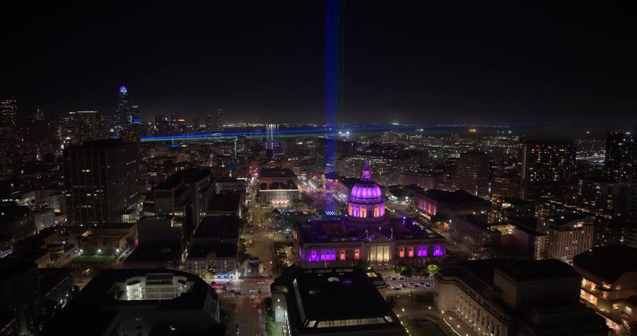 A slow aerial push-in shot of the iconic San Francisco City Hall at night, illuminated in purple with a striking blue beam of light rising behind it