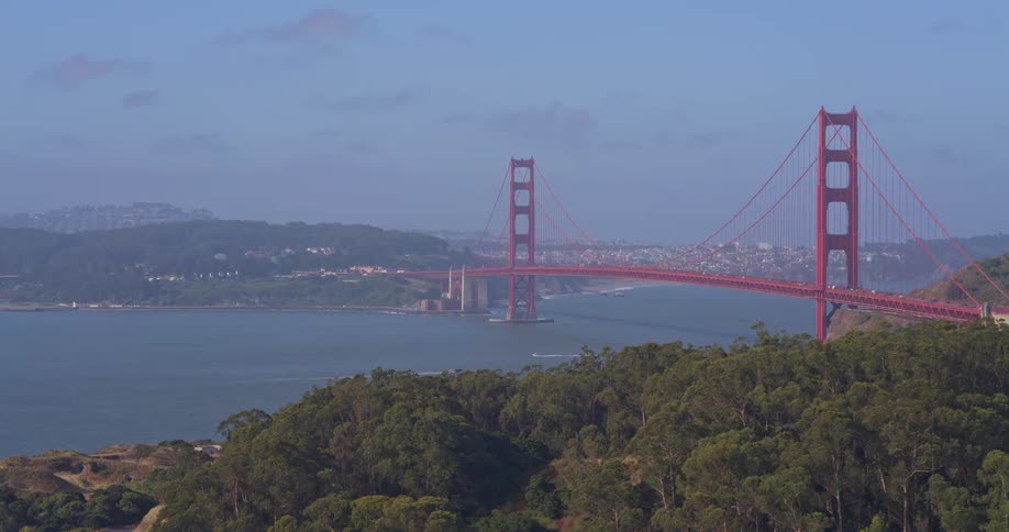A slow zoom-in aerial shot captures the iconic Golden Gate Bridge on a hazy day, with the San Francisco cityscape in the background and lush hills in the foreground