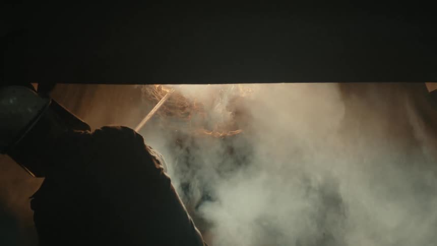 A silhouetted welder works in a smoky, dark workshop with sparks flying from the bright welding torch in slow motion