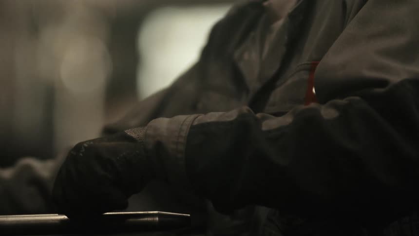 A close-up slow-motion shot of a worker's gloved hand resting on a metal pipe in a dimly lit industrial factory