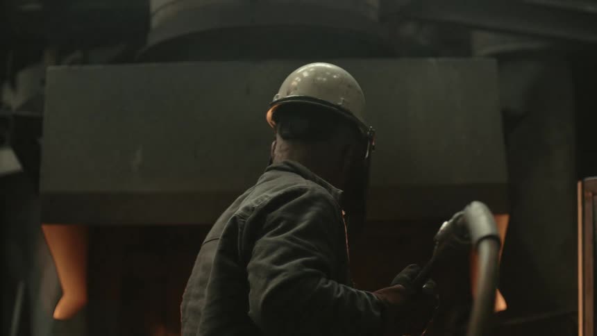 A foundry worker in a hard hat and protective gear works in front of a glowing hot furnace in slow motion