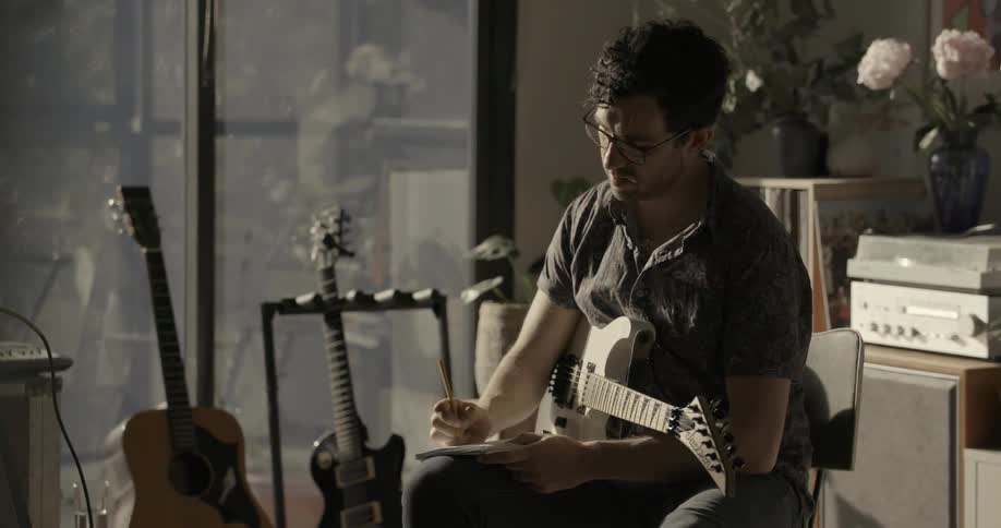 A man sits in a chair writing and holding a guitar in his lap