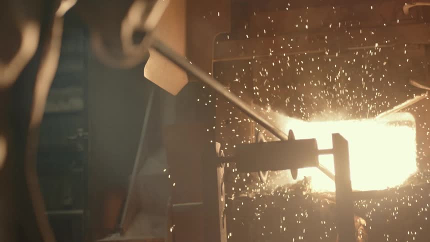 Slow motion footage shows a steelworker manipulating molten metal in a glowing furnace, causing bright sparks to erupt into the dark foundry