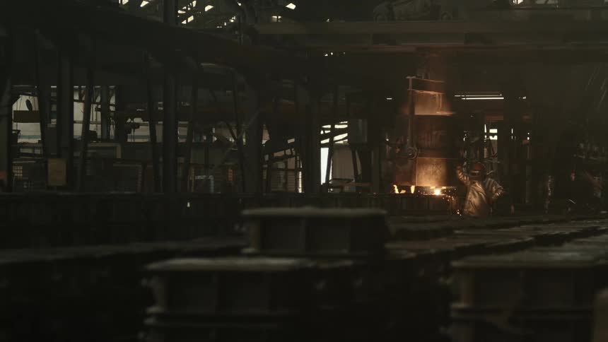 A worker in a full protective suit tends to a vat of molten metal in a dimly lit industrial factory