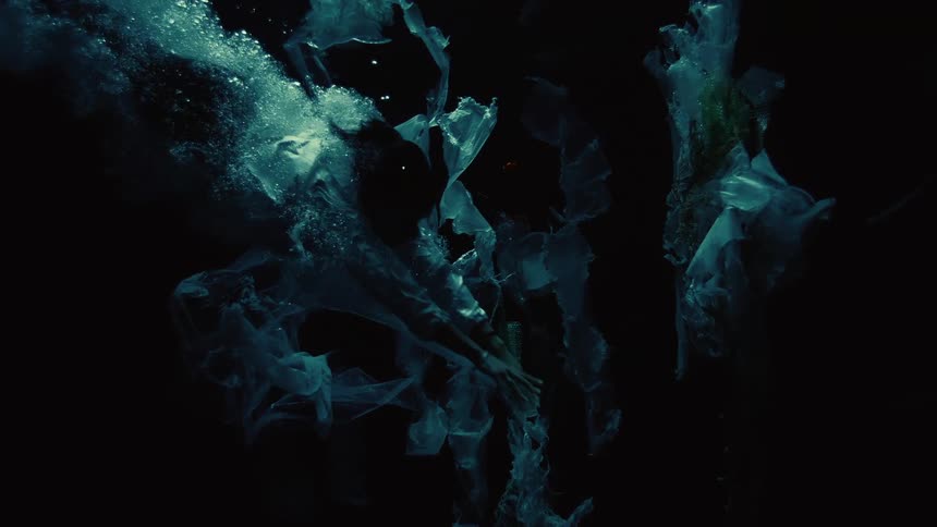 A dramatic underwater slow-motion shot shows a person trapped and sinking amidst plastic bags and trash