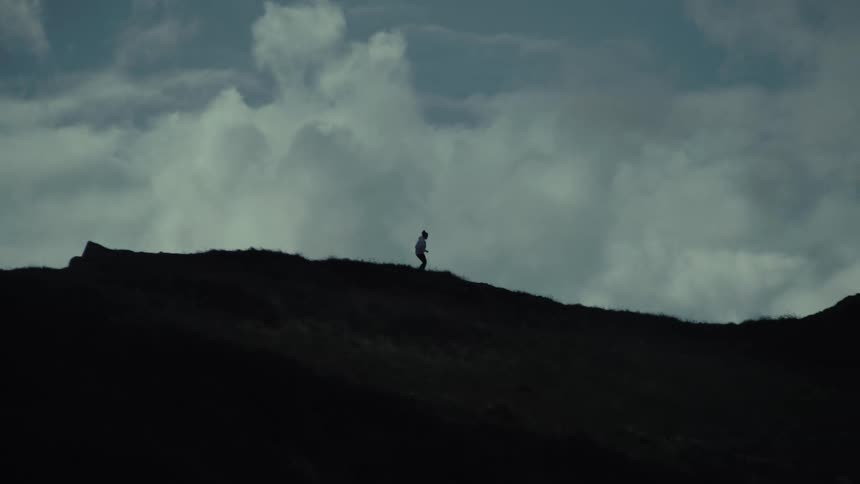 Silhouette of a person running along a grassy ridge against a cloudy sky
