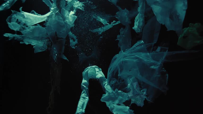 A person dressed in white sinks slowly through dark, polluted water, surrounded by a cloud of floating plastic bags