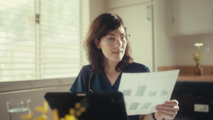 A female doctor in scrubs studies medical charts and MRI scans at her sunlit dining table while working from home