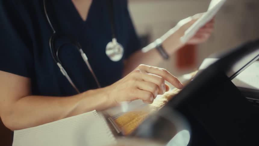 A doctor in scrubs with a stethoscope reviews medical documents while using a tablet