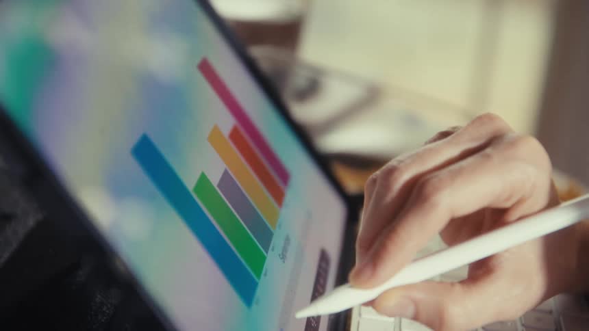 A close-up shot shows a hand using a digital pencil to analyze a colorful bar chart on a tablet