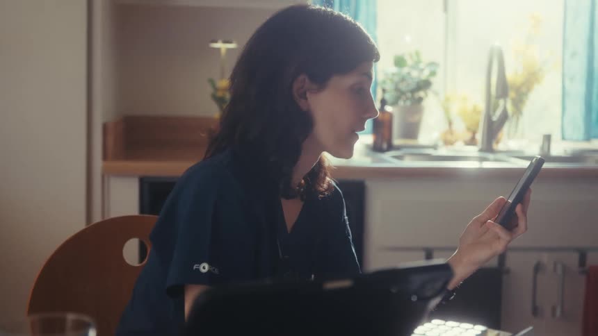 A woman in scrubs sits at her kitchen table and scrolls through her phone in the morning light