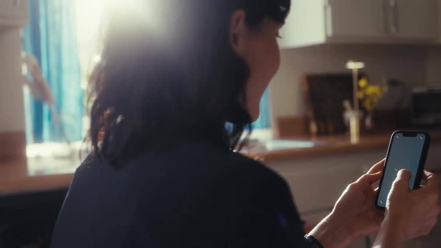Over-the-shoulder shot of a woman scrolling through her phone in a brightly lit kitchen