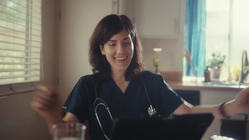 A smiling female doctor in scrubs joyfully celebrates good news she sees on her tablet at her sunlit kitchen table