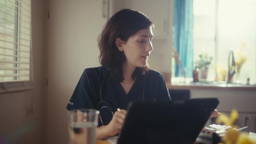 A woman in medical scrubs with a stethoscope studies on a tablet while sitting at her kitchen table at home