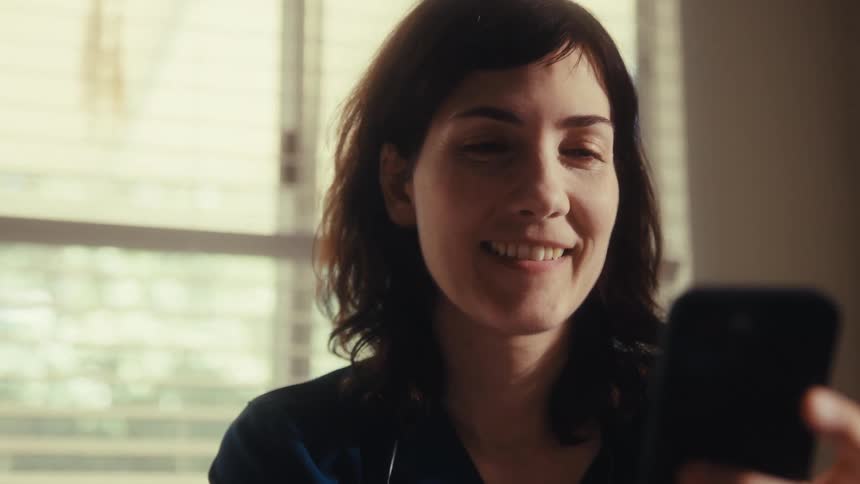 A female medical professional in scrubs smiles while using her smartphone during a break, with a slow zoom into her happy face