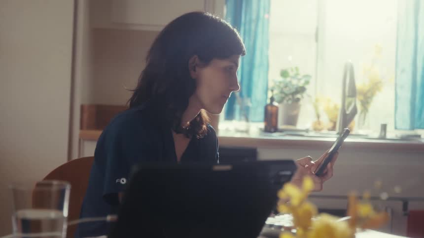 A female doctor in scrubs takes a break at her sunlit kitchen table while scrolling on her smartphone