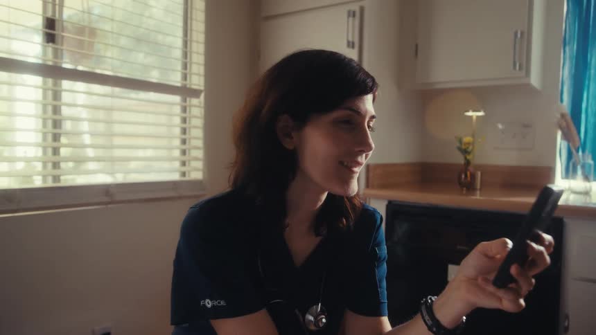 A female doctor in scrubs smiles warmly while looking at her smartphone, taking a well-deserved break at her sunlit kitchen table
