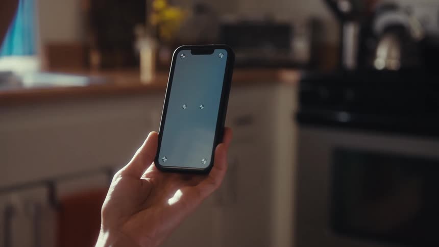 Over the shoulder shot of a person's hand holding a smartphone with tracking markers on the screen inside a sunlit kitchen