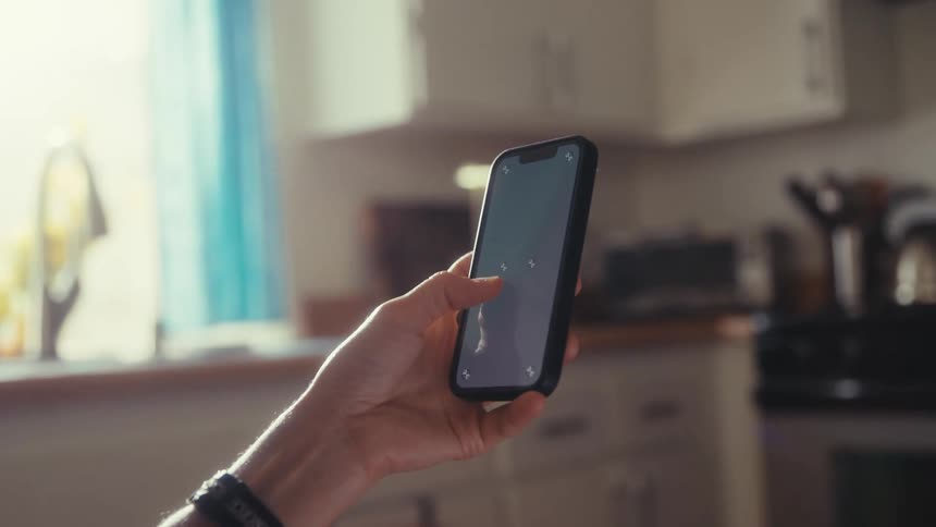 An over-the-shoulder view of a person holding a smartphone with tracking markers on the screen inside a sunlit kitchen