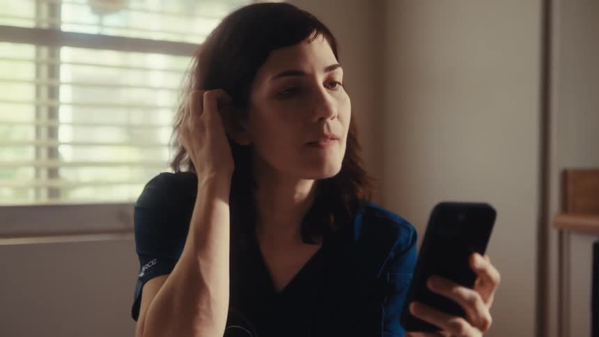 A tired female doctor in scrubs sits in her office and looks at her phone with a concerned expression as the camera slowly zooms in