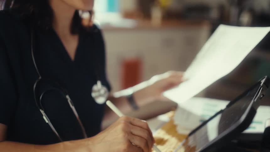 A close-up of a medical professional in scrubs using a stylus and finger on a tablet while reviewing paperwork