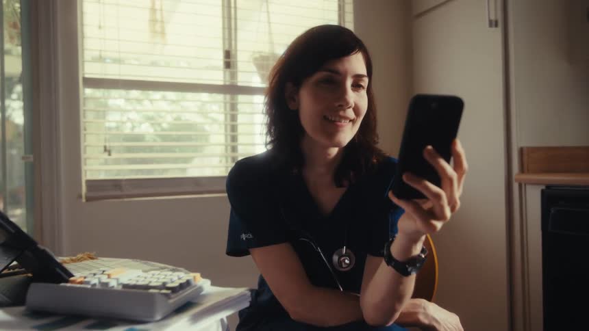A smiling female doctor in scrubs takes a break at her kitchen table and looks at her smartphone