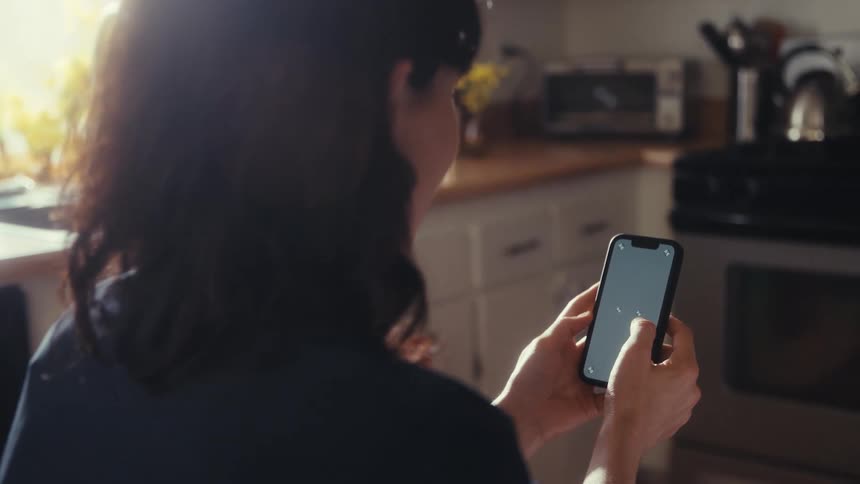 An over-the-shoulder shot of a woman scrolling on a smartphone with a trackable screen in a sunlit kitchen
