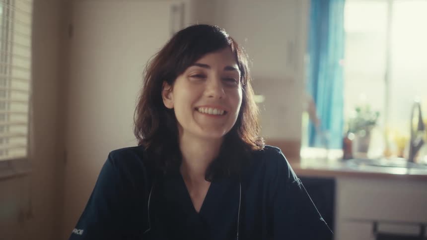 A happy female doctor in scrubs works from home on her tablet, smiling and laughing in her sunlit kitchen