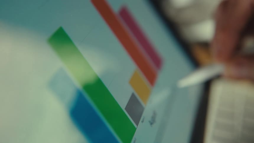 A financial analyst's hand uses a stylus to interact with a colorful digital bar graph on a tablet, analyzing business performance statistics