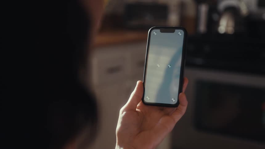 Close up over the shoulder shot of a person holding a smartphone with tracking markers on the screen inside a home kitchen