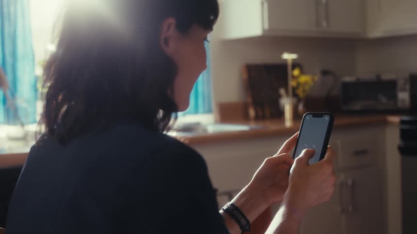 Over the shoulder shot of a woman scrolling on her smartphone in a brightly sunlit kitchen
