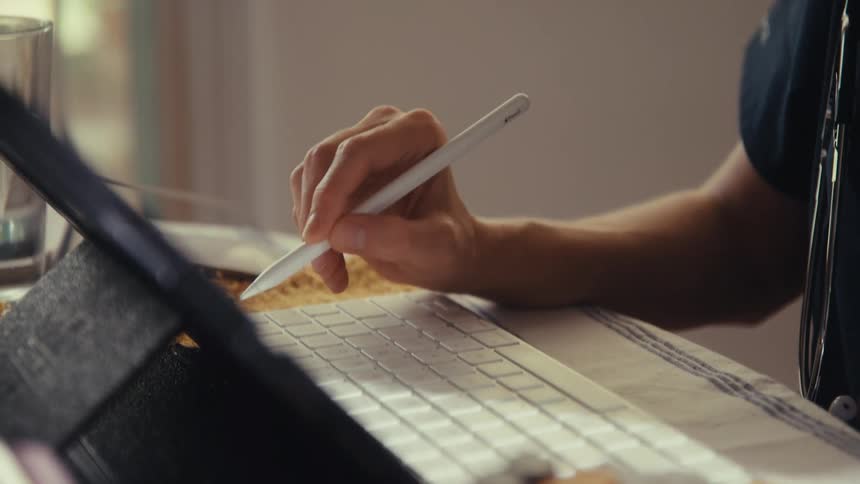 A medical professional wearing scrubs uses a digital stylus to work on a tablet at their desk