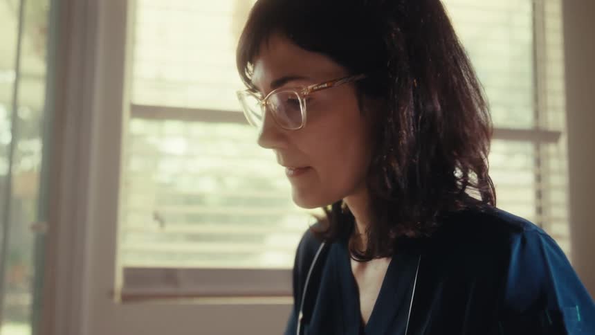A female doctor in glasses and scrubs calculates finances at her desk with a tablet and paperwork