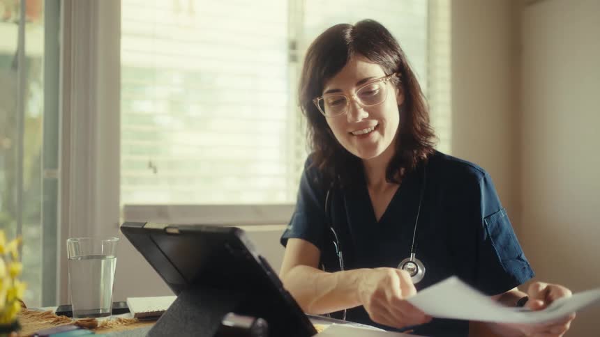 Slow motion shot of a smiling female doctor in scrubs working from home, reviewing documents at her sunlit kitchen table
