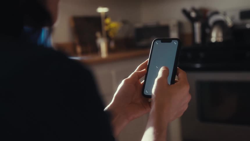 An over-the-shoulder shot of a person using a smartphone with a chroma key screen inside a sunlit kitchen