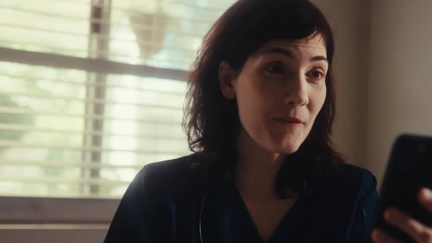 A smiling nurse in scrubs video chats on her phone at her kitchen table, her expression shifting to surprise and joy
