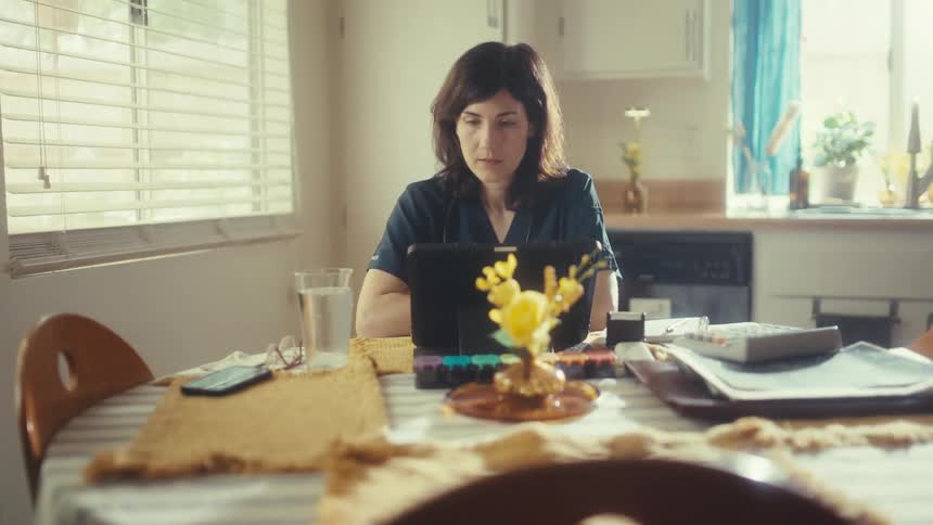 A female healthcare professional in scrubs works from home on her tablet at the sunlit kitchen table